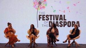 Four women converse on a stage while sitting in white chairs and holding microphones. Behind them is a white backdrop with the Festival of the Diaspora logo.
