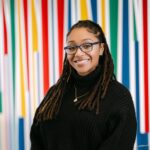 This photograph shows a woman standing in front of colorful wall. It's a headshot of the woman.